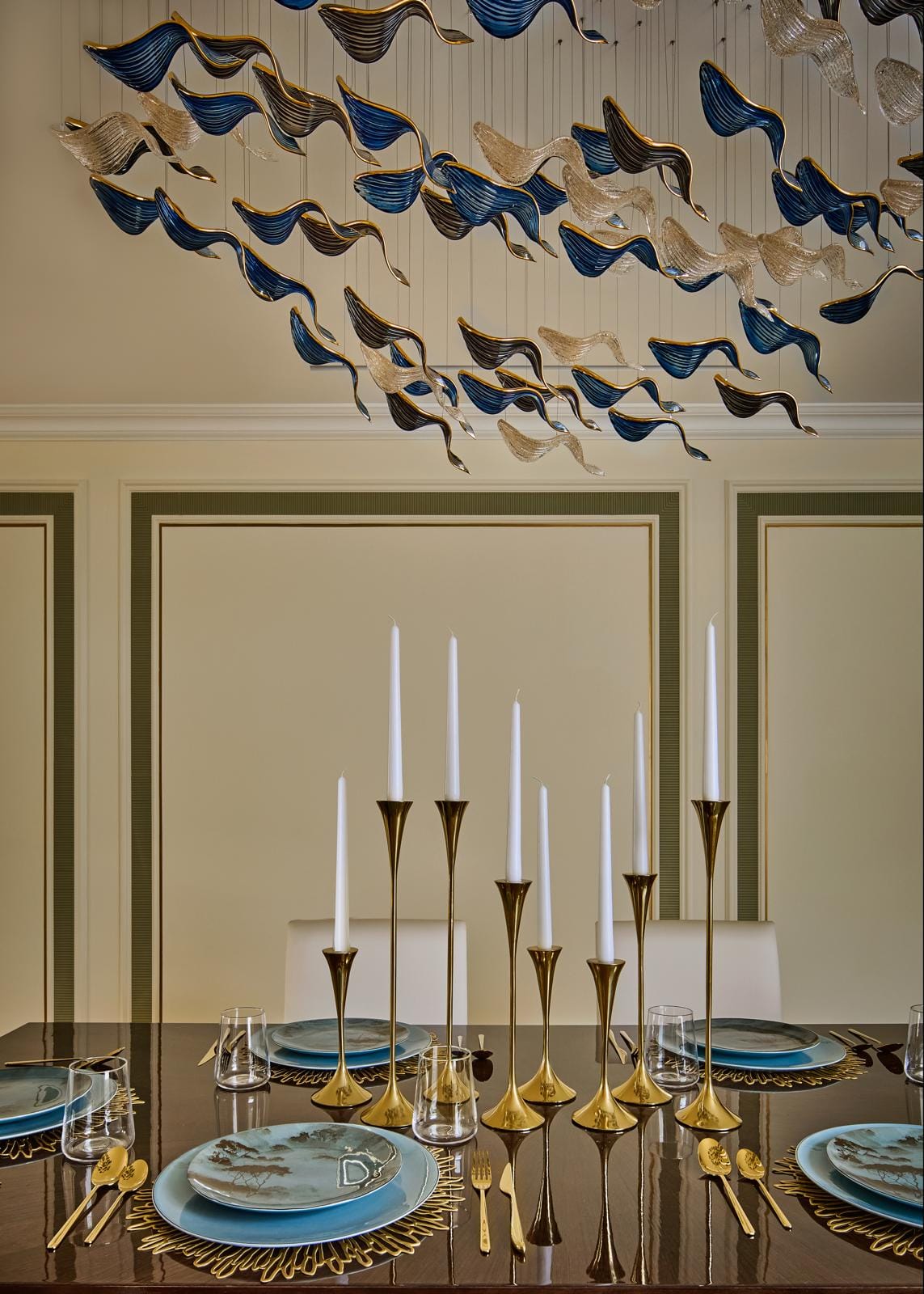 Dining table dressed with brass candlesticks under a blue glass ceiling artwork.